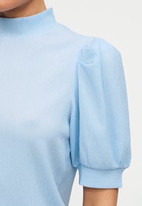 Ανοιχτό μπλε υφασμάτινο κοντομάνικο τοπ με φουσκωτά μανίκια και ψηλό στρογγυλό λαιμόκοψη σε ένα άτομο με λευκό απλό φόντο.