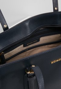 Laivastonsininen synteettisestä nahasta valmistettu käsilaukku vetoketjullisella osastolla, kullanvärisillä metalliosilla ja teksturoidulla ulkopinnalla. Sisäpuolella on brändin logokuvioitu vuori.