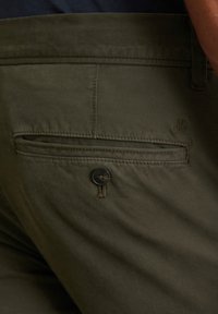 Olivgrüne Hose mit glatter Textur, ausgestattet mit einer Gesäßtasche, die mit einem schwarzen Knopf gesichert ist, sowie einer dezenten Logo-Prägung.