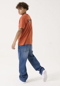 Oranges Baumwoll-T-Shirt mit schwarzem Text auf dem Rücken, kombiniert mit lockeren blauen Jeans und weißen Sneakers.