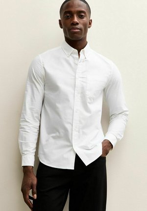 Man staat tegen een muur, draagt een wit overhemd met knoopsluiting en zakje, één hand in de zwarte broekzak, neutrale uitdrukking, kijkt recht vooruit.