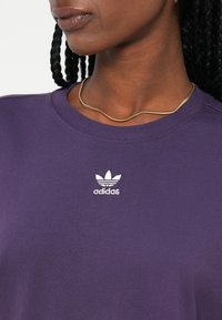 Violetti puuvillapaita, jossa on valkoinen adidas-logo rinnassa, pyöreä pääntie ja lyhyet hihat. Sileä pinta ja klassinen istuvuus.