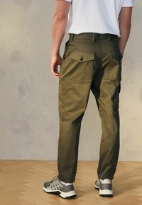 Olivenfarvede cargo-bukser med tapered fit, der har to baglommer med klapper og en glat stofoverflade. Båret med grå atletiske sko.