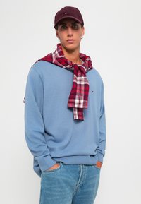 Maglione blu chiaro con polsini a coste, indossato sopra una sciarpa a scacchi rossi e bianchi. Un cappello bordeaux completa l'outfit casual.