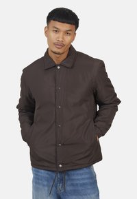 Veste matelassée marron avec col, boutons-pression et poches latérales ; possède une texture lisse et des poignets élastiques ; portée sur un jean bleu.