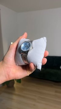 Silver wristwatch with a blue dial, stainless steel bracelet, displayed on a gray cushion in a hand, against a blurred background.