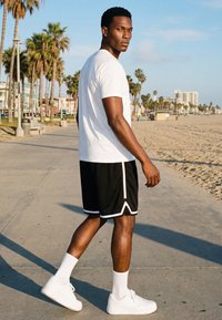 Joven caminando por un sendero junto a la playa, vistiendo una camiseta blanca, pantalones cortos negros, calcetines blancos y zapatillas blancas, con palmeras y edificios de fondo.