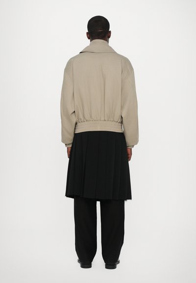Beige Jacke mit hohem Kragen und elastischem Bund, kombiniert mit einem schwarzen Faltenrock und lockeren schwarzer Hose, von hinten gesehen.
