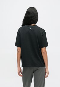 Άτομο με μακριά μαύρα μαλλιά που φαίνεται από πίσω, φορώντας ένα μαύρο μπλουζάκι της Jordan και γκρίζα παντελόνια, σε απλό λευκό φόντο.