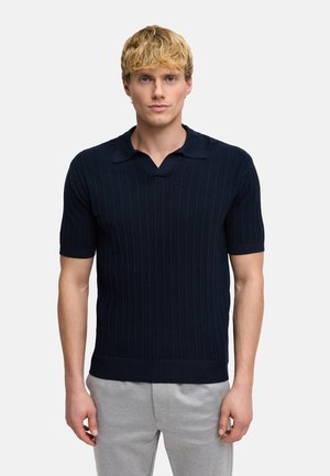 Junger Mann mit kurzen blonden Haaren, trägt ein marineblaues, kurzärmeliges, geripptes Poloshirt und hellgraue Hosen vor weißem Hintergrund.