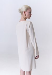 Helles cremefarbenes Kleid mit langen Ärmeln, geradem Schnitt und glatter Textur. Der Rücken hat einen weiten Ausschnitt und dezente Details.