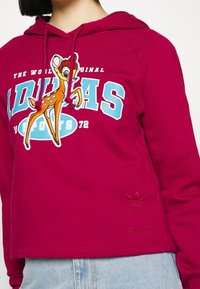 Burgundy Adidas hoodie with blue and white logo, Bambi graphic on chest, Adidas and Disney logos embroidered near hem, worn with light blue jeans.