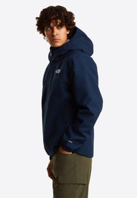 Veste imperméable marine avec capuche et poignets élastiques, arborant un logo sur la poitrine. Associée à un pantalon vert olive contrastant.