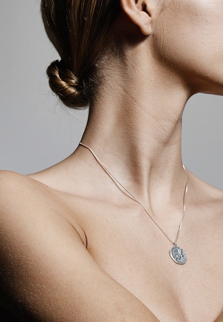 Collier en argent avec un pendentif ovale présentant des détails complexes, porté autour d'un cou nu sur un fond neutre.