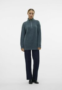 Temno teal pulover s visokim ovratnikom in pol-zadrgo, ohlapen kroj, strukturirano pletenje, v kombinaciji s temno denim hlačami ravne kroja in črnimi gležnjački.