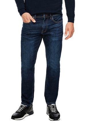 Hombre con jeans ajustados azul oscuro, zapatillas negras y blancas, y una camiseta negra de manga larga, con una mano en el bolsillo.