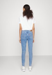 Jeans ajustados de color azul claro con una altura media, que presentan dos bolsillos traseros, una etiqueta de cuero marrón y detalles de costura. Llevados con una camiseta blanca.