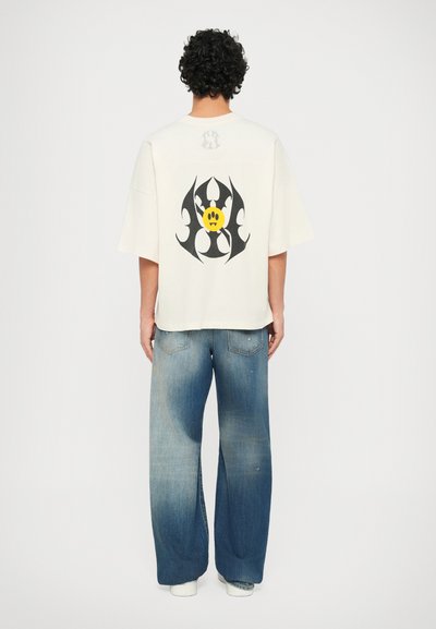 Oversized cream cotton t-shirt with a black and yellow graphic design on the back, paired with loose blue denim jeans.