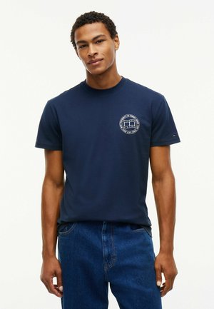 Junger Mann trägt ein dunkelblaues T-Shirt mit einem kleinen runden Logo auf der linken Brust und blaue Jeans und steht vor einem weißen Hintergrund.