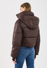 Ruda pūkinė striukė su aukštu apykakle ir kapišonu, su quilted dizainu ir išriškomis rankovėmis. Dėvima su mėlynomis džinsinėmis kelnėmis.