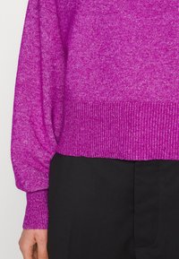 Pull en tricot fuchsia avec un ourlet côtelé et des poignets de manches, présentant une texture douce. Associé à un pantalon noir taillé sur mesure.