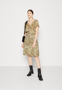 Beiges Druckkleid mit braunen Spritzern, kurzen Ärmeln, V-Ausschnitt und Taillengürtel. Trägt dazu schwarze Stiefeletten und eine kleine schwarze Tasche.