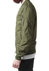 Veste bomber vert olive avec une texture lisse, des poignets et un ourlet côtelés, et une poche zippée sur le bras gauche.