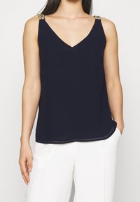 Marinblå linne med V-ringning och metallkedjeprydnader på axelremmarna. Tillverkat av ett mjukt tyg; avkopplad passform.