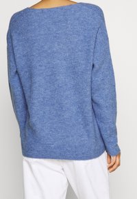 Vue arrière d'une personne portant un pull bleu doux associé à un pantalon blanc, mettant en valeur la texture du tissu et la coupe décontractée.