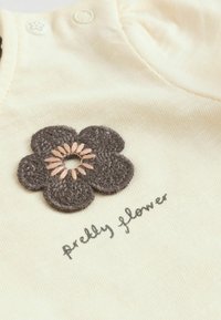 Tissu crème avec une fleur crochetée gris foncé et "jolie fleur" écrit à la main en dessous en écriture grise.