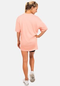 Overmaat perzikt-shirt met korte mouwen, ronde halslijn en een gladde textuur. Gekenmerkt door het dragen van zwarte shorts en witte sportschoenen.