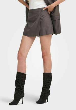 Femme portant une mini-jupe plissée marron, un haut blanc et des bottes noires souples à talons hauts, debout avec une main dans la poche de la jupe.