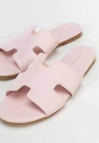 Paar lichtroze platte slides met brede H-vormige bandjes en beige zolen op een witte achtergrond.