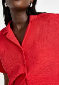 Camicia rossa a maniche corte con texture, colletto e chiusura frontale a bottoni. Tessuto morbido con un sottile motivo stropicciato.