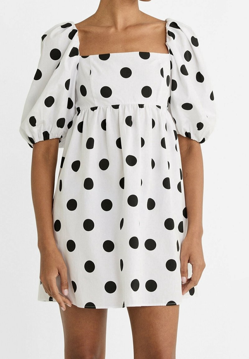Vestido blanco con estampado de lunares negros, escote cuadrado y mangas abullonadas. Hecho de tela liviana con un diseño de falda acampanada.