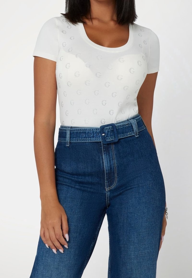 Femme portant un haut blanc à manches courtes avec un motif subtil, associé à un jean bleu foncé taille haute et une ceinture en denim assortie.