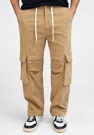 Uomo con pantaloni cargo beige con molte tasche, coulisse bianca, scarpe da ginnastica bianche e nere, mani infilate nelle tasche, in piedi su un pavimento bianco.