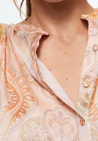 Blusa de un suave tono rosa claro con un delicado patrón de cachemira en naranja y crema, con un frontal abotonado y detalle en el cuello. Textura de tela suave.