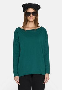 Lniany top w kolorze teal z długim rękawem i szerokim dekoltem, wykonany z miękkiego materiału. Noszony z czarną, szydełkowaną czapką w kształcie uszu kota oraz dużymi okularami przeciwsłonecznymi.