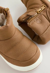 Hnedé ankle boots s textúrovaným syntetickým materiálom, prešívanými detailmi a bielou gumovou podrážkou. Obsahujú bočný zips pre jednoduché obúvanie.
