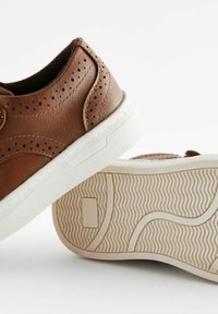 Baskets en cuir marron featuring un design perforé, semelle en caoutchouc blanche et semelle texturée avec des motifs ondulés.