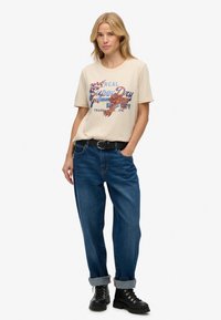 Beżowa bawełniana koszulka z nadrukiem w niebieskim i czerwonym kolorze, w połączeniu z niebieskimi dżinsami, podwiniętymi mankietami i czarnymi sznurowanymi butami.