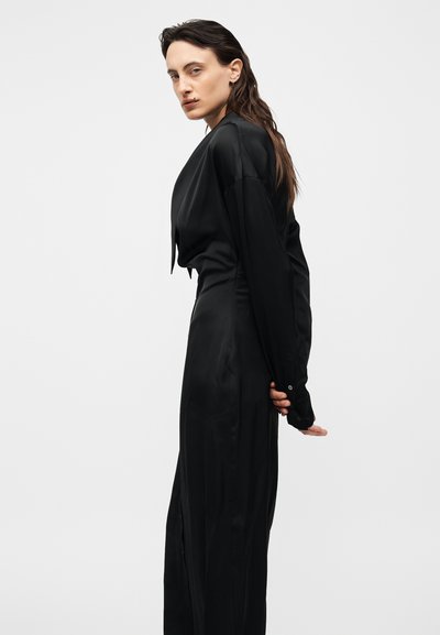 Γυναίκα με μακριά μαλλιά που φορεί μαύρο σατέν φόρεμα με μακριά μανίκια, στέκεται πλάγια με τα χέρια πιεσμένα πίσω από την πλάτη μπροστά σε μια απλή φόντο.