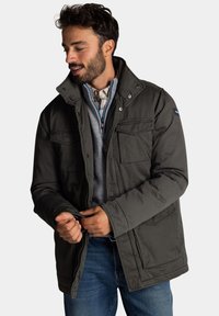 Grijze geïsoleerde jas met een hoge kraag, meerdere voorzakken en een rits. Draagt over een lichtblauw sweater en een geruit overhemd.