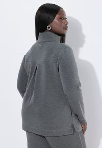 Haut gris à manches longues avec col montant, fentes latérales et un pli discret à l'arrière, fabriqué dans un tissu doux à la texture lisse.