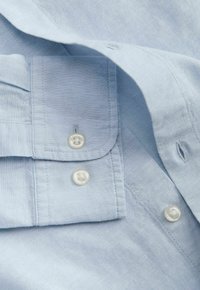 Chemise bleu clair à manches longues avec poignets boutonnés et boutons blancs visibles sur une texture de tissu douce.