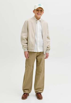 Dreng smilende iført beige jakke, hvid skjorte, khakifarvede bukser, brune sko og hvid baseballkasket på ensfarvet lyst baggrund.