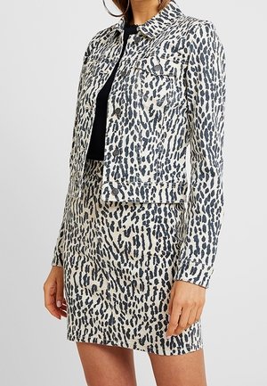 Vrouw draagt een zwart-wit gevlekte jas en een bijpassende rok over een zwart shirt, met zichtbare oorringen.