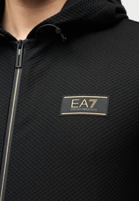 Μαύρο φούτερ με φερμουάρ, με υφή υφάσματος, διαθέτοντας μπροστινό λογότυπο σε χρυσό και μαύρο και κορδόνια στο καπό.