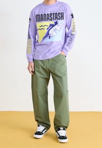 Osoba w fioletowej bluzie z długim rękawem, na której widnieje kolorowy napis "MANASTASH", w oliwkowych spodniach i czarno-białych sneakersach, stoi na żółtej podłodze.